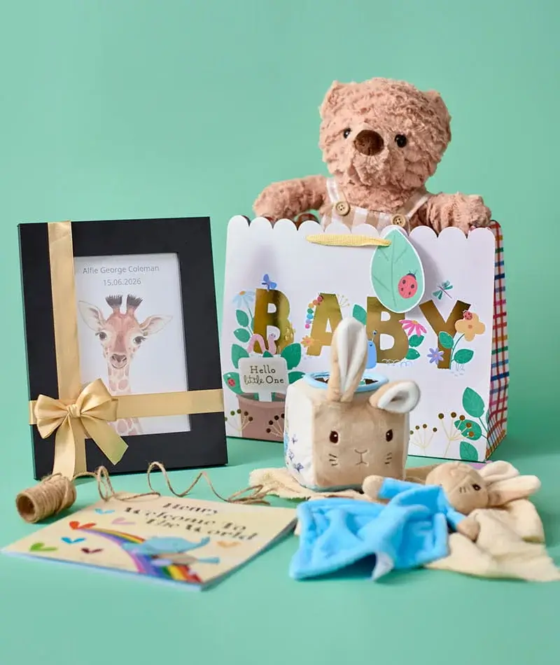 A baby gift display on a mint green background featuring a teddy bear in a decorative baby gift bag, a framed personalised print, soft plush toys, a blanket, and keepsake cards.